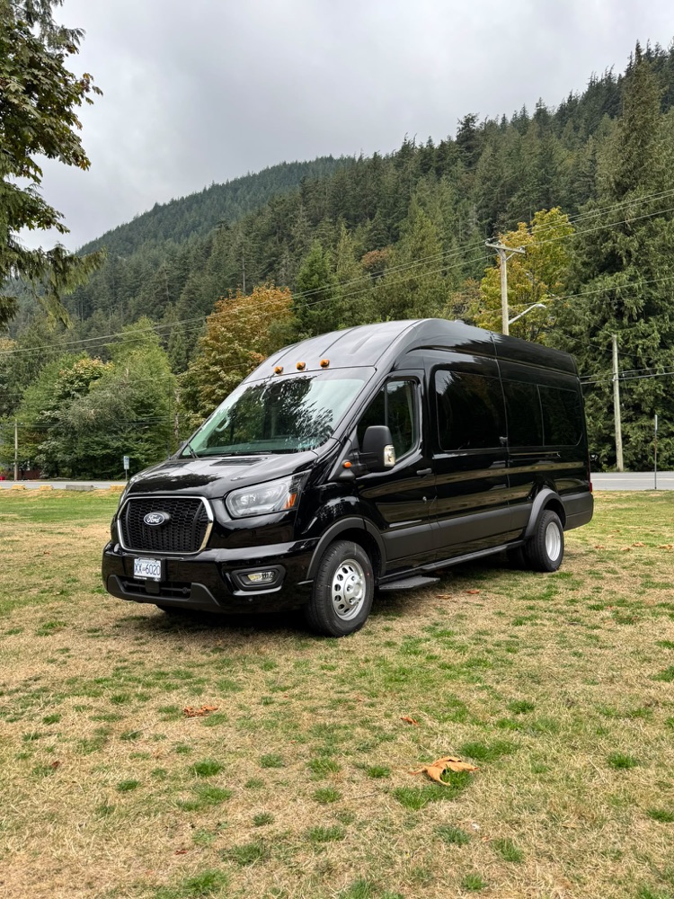 Black Ford Transit Wagon XLT 2026, front view in a park with forested mountains behind